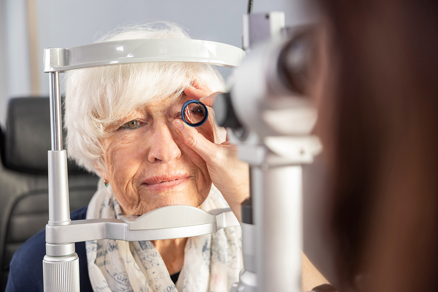 Augenuntersuchung bei Macuria Eine ältere Frau unterzieht sich einer Augenuntersuchung mit einem Gerät, das auf das Auge gerichtet wird, während eine Fachkraft hilft.